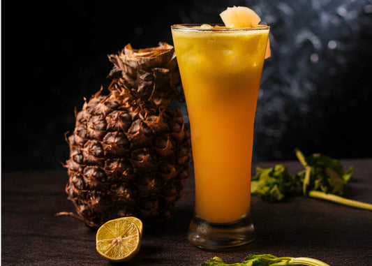 A tall glass of vibrant yellow pineapple martini juice garnished with a pineapple wedge, surrounded by a fresh whole pineapple, a sliced lime, and sprigs of green herbs, set against a dark background for a tropical and refreshing look.