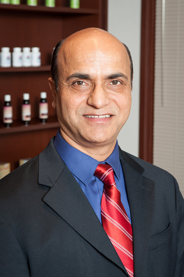 Dr. Shailinder Sodhi in a suit and tie stands confidently before shelves filled with various medicines in a well-organized setting.