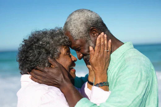 An older couple shares a warm embrace on the beach, surrounded by gentle waves and a serene sunset backdrop.