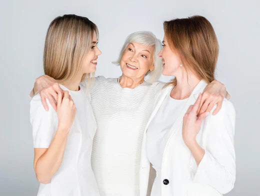 Three women in white attire stand together with an elderly woman, showcasing a moment of unity and warmth among generations.